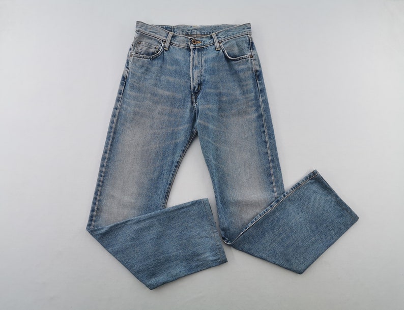 May include: Light blue denim jeans with a classic five-pocket design. The jeans have a button fly and a faded wash, giving them a vintage look. The jeans are laid flat on a light gray surface.