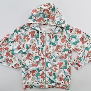 May include: White zip-up hoodie featuring a repeating pattern of a red-haired mermaid with a green tail. The hoodie has a drawstring hood and a full-length zipper. The design includes other sea creatures and plants.