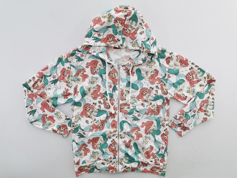 May include: White zip-up hoodie featuring a repeating pattern of a red-haired mermaid with a green tail. The hoodie has a drawstring hood and a full-length zipper. The design includes other sea creatures and plants.