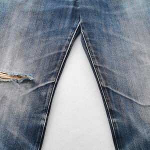 K&ouml;nnte beinhalten: Nahaufnahme einer blauen Jeans mit einem Used-Look. Die Jeans hat ein gro&szlig;es, ausgefranstes Loch am linken Knie. Der Stoff weist Ausbleichungen und Abnutzungsmuster auf, typisch f&uuml;r Vintage-Denim.