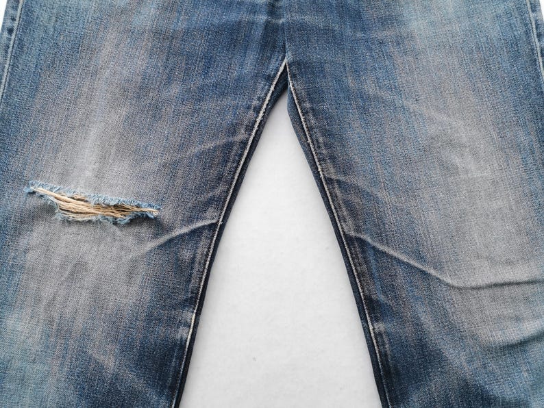 K&ouml;nnte beinhalten: Nahaufnahme einer blauen Jeans mit einem Used-Look. Die Jeans hat ein gro&szlig;es, ausgefranstes Loch am linken Knie. Der Stoff weist Ausbleichungen und Abnutzungsmuster auf, typisch f&uuml;r Vintage-Denim.