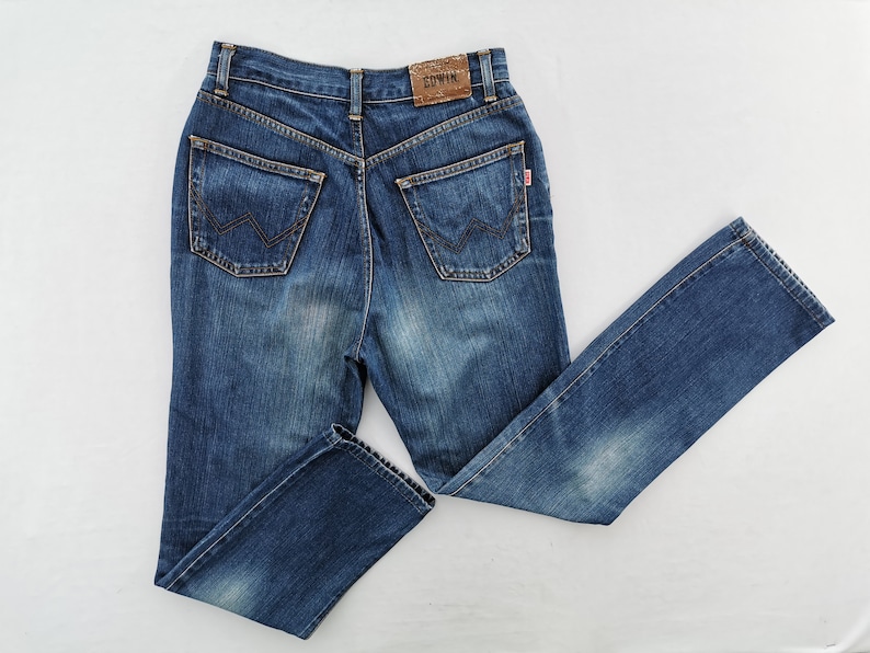 May include: Blue denim jeans with a classic five-pocket design. Features a high waist and straight leg. The back pockets have a distinctive 'W' stitch. A leather patch and a red tag are visible. The jeans are laid flat on a white surface.