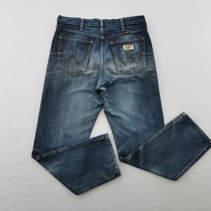 K&ouml;nnte beinhalten: Blaue Jeans aus Denim mit klassischem F&uuml;nf-Taschen-Design. Die Jeans haben eine verwaschene Optik und einen Wrangler-Markenaufn&auml;her auf der Ges&auml;&szlig;tasche. Die Jeans liegen flach auf einer wei&szlig;en Oberfl&auml;che.