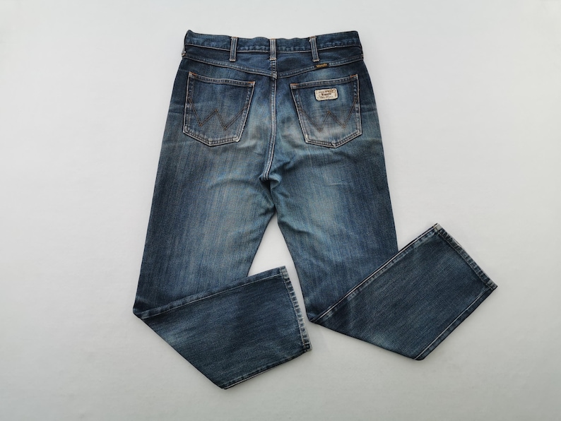K&ouml;nnte beinhalten: Blaue Jeans aus Denim mit klassischem F&uuml;nf-Taschen-Design. Die Jeans haben eine verwaschene Optik und einen Wrangler-Markenaufn&auml;her auf der Ges&auml;&szlig;tasche. Die Jeans liegen flach auf einer wei&szlig;en Oberfl&auml;che.