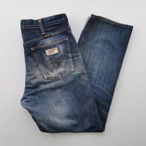 K&ouml;nnte beinhalten: Blaue Wrangler Jeans aus Denim mit verwaschenem Look. Die Jeans haben ein klassisches F&uuml;nf-Taschen-Design, einen Markenaufn&auml;her auf der Ges&auml;&szlig;tasche und ein Wrangler-Logo. Die Jeans sind gefaltet und flach hingelegt.
