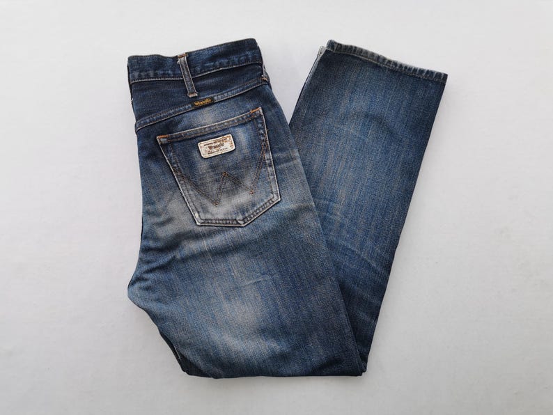 K&ouml;nnte beinhalten: Blaue Wrangler Jeans aus Denim mit verwaschenem Look. Die Jeans haben ein klassisches F&uuml;nf-Taschen-Design, einen Markenaufn&auml;her auf der Ges&auml;&szlig;tasche und ein Wrangler-Logo. Die Jeans sind gefaltet und flach hingelegt.
