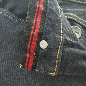 K&ouml;nnte beinhalten: Nahaufnahme einer dunklen Jeans. Die Jeans hat ein schwarz-rotes Streifendetail am Bund und goldfarbene N&auml;hte. Ein silberner Knopf ist sichtbar. Die Gesamt&auml;sthetik ist l&auml;ssig und stilvoll.