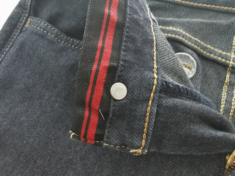 K&ouml;nnte beinhalten: Nahaufnahme einer dunklen Jeans. Die Jeans hat ein schwarz-rotes Streifendetail am Bund und goldfarbene N&auml;hte. Ein silberner Knopf ist sichtbar. Die Gesamt&auml;sthetik ist l&auml;ssig und stilvoll.