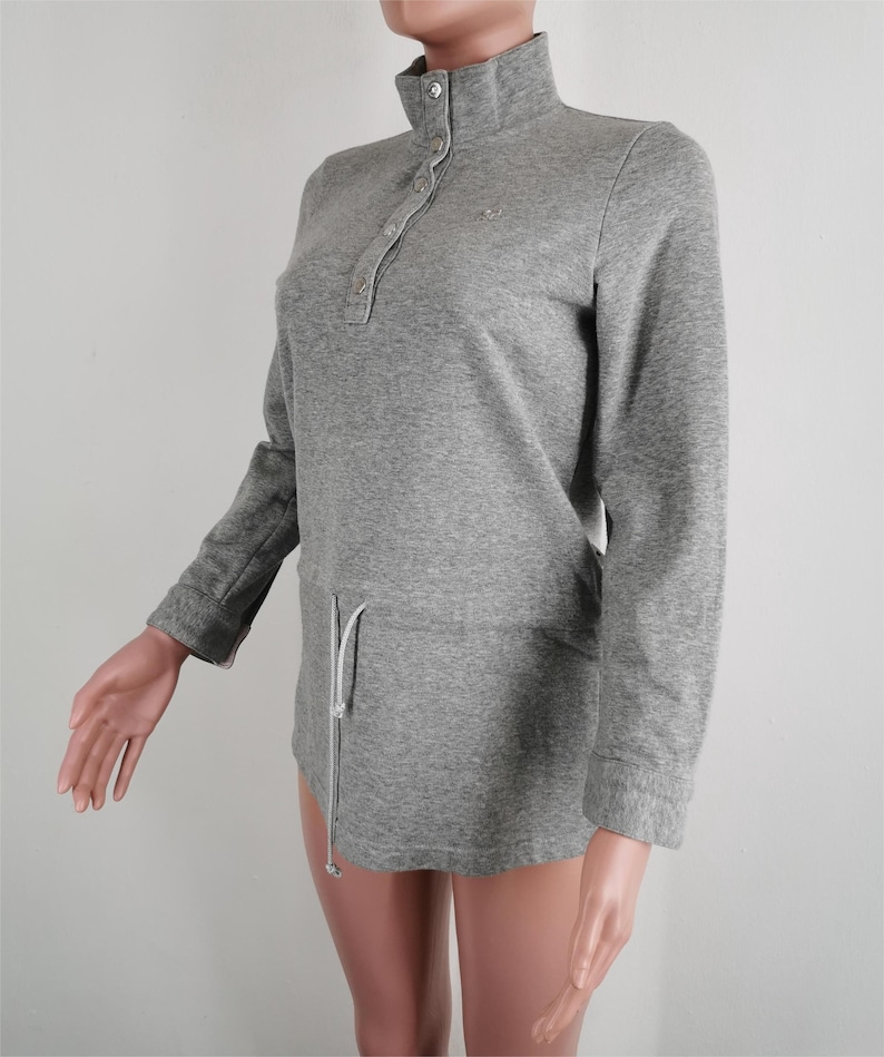 May include: A gray long-sleeve pullover shirt with a high collar and a button-down placket. The shirt has a drawstring waist and a small logo on the chest. The sleeves are long and the fabric appears to be a soft knit.