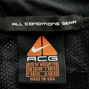 Pu&ograve; includere: Etichetta nera per indumenti con testo bianco "ALL CONDITIONS GEAR". L'etichetta presenta il logo Nike ACG, informazioni sulle taglie e "MADE IN USA". Il logo &egrave; un triangolo arancione con uno swoosh Nike bianco.