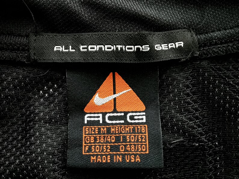 Pu&ograve; includere: Etichetta nera per indumenti con testo bianco "ALL CONDITIONS GEAR". L'etichetta presenta il logo Nike ACG, informazioni sulle taglie e "MADE IN USA". Il logo &egrave; un triangolo arancione con uno swoosh Nike bianco.