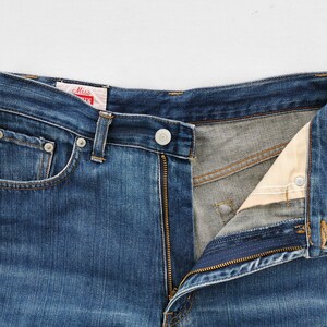 May include: Close-up of blue denim jeans with a partially unzipped fly. The jeans have a button closure, belt loops, and front pockets. A red and white tag is visible near the waistband. The jeans are a classic style.