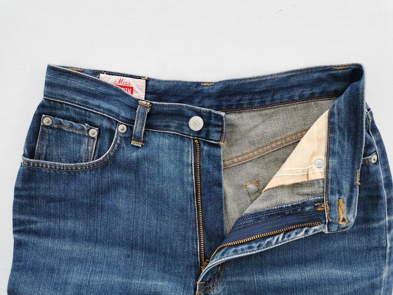 May include: Close-up of blue denim jeans with a partially unzipped fly. The jeans have a button closure, belt loops, and front pockets. A red and white tag is visible near the waistband. The jeans are a classic style.