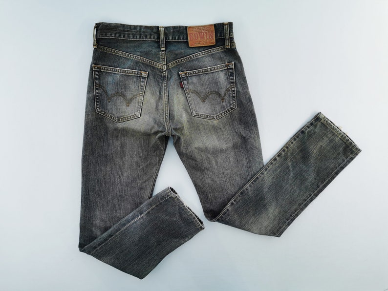 May include: Faded black denim jeans with a classic five-pocket design. The jeans feature a leather patch on the back waistband and a small red tag on the back pocket. The jeans are laid flat on a light blue surface.