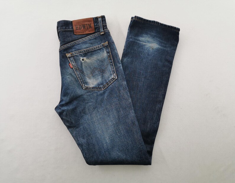 K&ouml;nnte beinhalten: Dunkelblaue Jeans mit verwaschenem Effekt und Used-Details. Die Jeans haben ein klassisches F&uuml;nf-Taschen-Design, einen Lederaufn&auml;her mit Markenname und ein rotes Logo-Etikett. Die Jeans liegen flach auf einer wei&szlig;en Oberfl&auml;che.