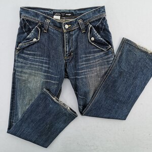 Puede incluir: Vaqueros de mezclilla azul con un lavado descolorido y detalles desgastados. Incluye dise&ntilde;os de bolsillos &uacute;nicos con cierres de bot&oacute;n y bragueta cl&aacute;sica con bot&oacute;n y cremallera. La etiqueta de la marca Wrangler es visible en la cintura.