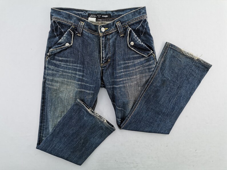 Puede incluir: Vaqueros de mezclilla azul con un lavado descolorido y detalles desgastados. Incluye dise&ntilde;os de bolsillos &uacute;nicos con cierres de bot&oacute;n y bragueta cl&aacute;sica con bot&oacute;n y cremallera. La etiqueta de la marca Wrangler es visible en la cintura.