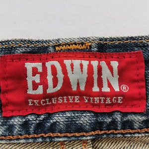 Puede incluir: Primer plano de una etiqueta roja con la marca "EDWIN" en blanco, sobre "EXCLUSIVE VINTAGE" en unos vaqueros azules. Los vaqueros tienen costuras visibles y un aspecto descolorido.