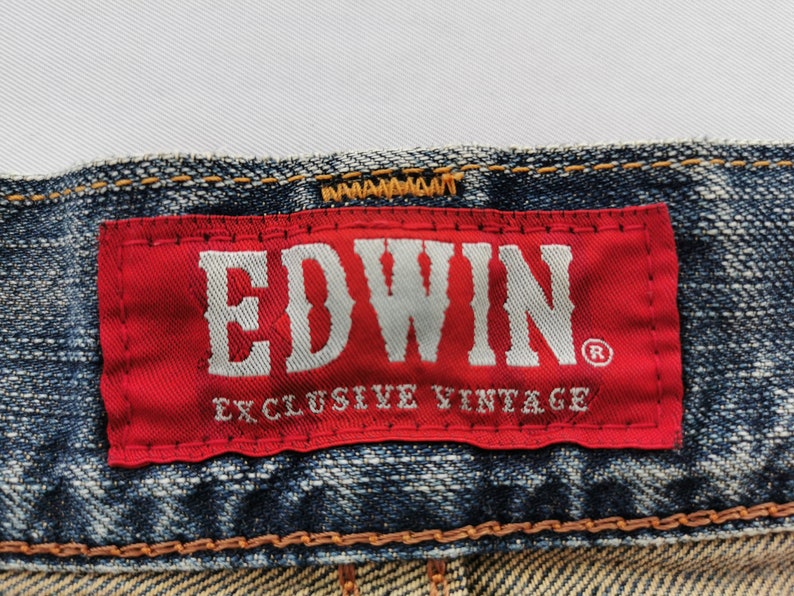 Puede incluir: Primer plano de una etiqueta roja con la marca "EDWIN" en blanco, sobre "EXCLUSIVE VINTAGE" en unos vaqueros azules. Los vaqueros tienen costuras visibles y un aspecto descolorido.