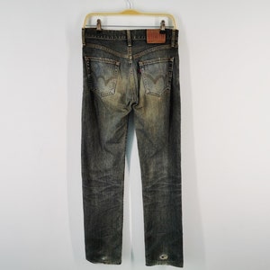 May include: Faded black denim jeans with a worn appearance, featuring a leather patch and the Levi's logo. The jeans are hanging on a gold hanger against a white background. The back pockets have the Levi's stitching.