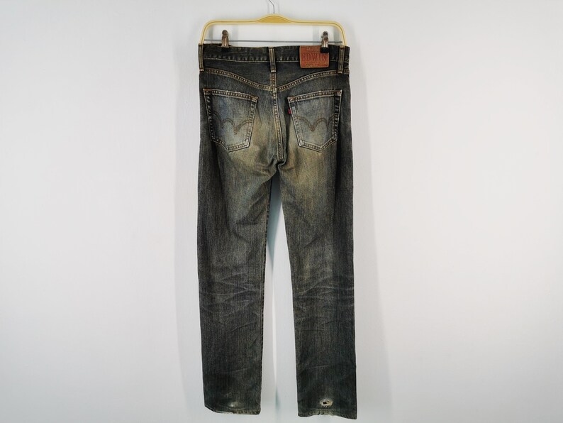 May include: Faded black denim jeans with a worn appearance, featuring a leather patch and the Levi's logo. The jeans are hanging on a gold hanger against a white background. The back pockets have the Levi's stitching.