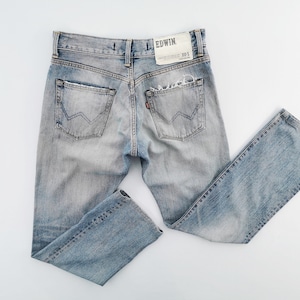 May include: Light blue denim jeans with distressed details, including a ripped pocket and faded areas. The back pockets have a distinctive W-shaped stitching. The Edwin brand label is visible. A classic, casual style.