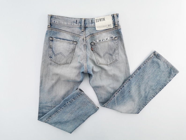 May include: Light blue denim jeans with distressed details, including a ripped pocket and faded areas. The back pockets have a distinctive W-shaped stitching. The Edwin brand label is visible. A classic, casual style.