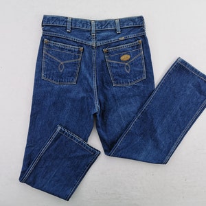 May include: Dark blue denim jeans with a classic five-pocket design. Features include a button and zipper closure, belt loops, and two back pockets with decorative stitching. The jeans are a straight-leg style.