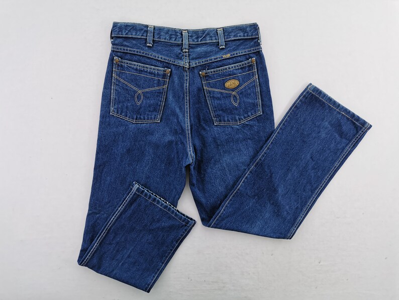 May include: Dark blue denim jeans with a classic five-pocket design. Features include a button and zipper closure, belt loops, and two back pockets with decorative stitching. The jeans are a straight-leg style.