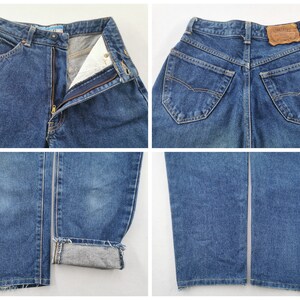 May include: Blue denim jeans with a classic five-pocket design. The jeans feature a zip fly, button closure, and a leather patch on the back. The cuffs are rolled up, revealing the interior fabric. The jeans are a medium wash.