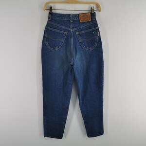 May include: Dark wash denim jeans with a high waist and a classic five-pocket design. The jeans are hanging on a wooden hanger. A leather patch is visible on the back waistband. The jeans have a straight leg cut.
