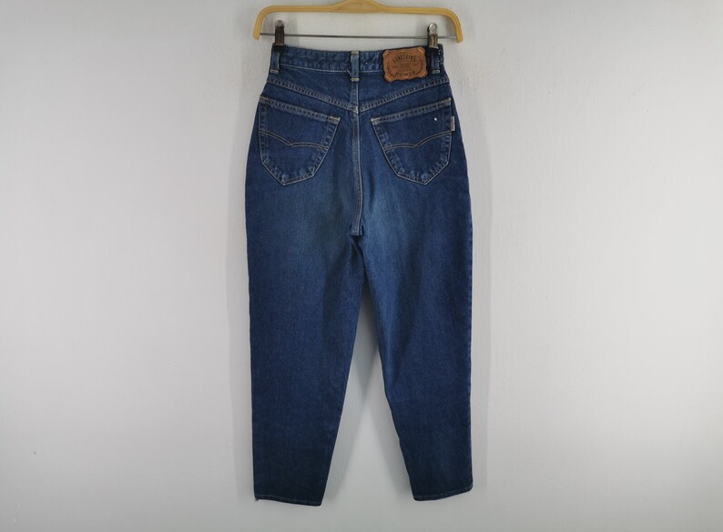 May include: Dark wash denim jeans with a high waist and a classic five-pocket design. The jeans are hanging on a wooden hanger. A leather patch is visible on the back waistband. The jeans have a straight leg cut.