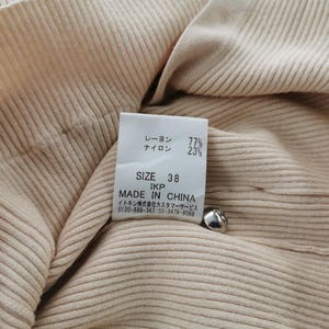 May include: Close-up of a beige ribbed knit fabric with a label. The label indicates the fabric composition: 77% rayon, 23% nylon, and size 38. The label also states "MADE IN CHINA". A silver button is visible.