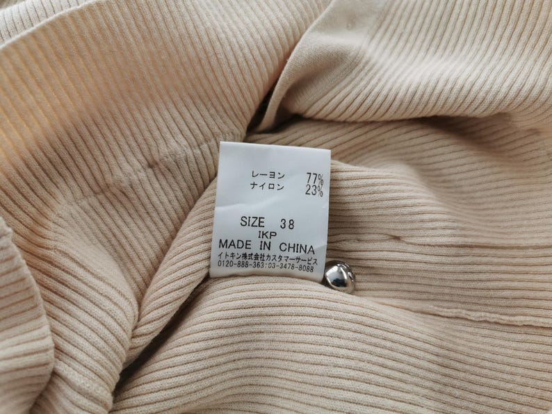 May include: Close-up of a beige ribbed knit fabric with a label. The label indicates the fabric composition: 77% rayon, 23% nylon, and size 38. The label also states "MADE IN CHINA". A silver button is visible.