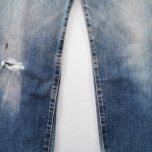 May include: Close-up of blue denim jeans with a distressed, worn appearance. The jeans have a ripped section on the left leg, showing frayed edges. Fading and discoloration are visible, adding to the vintage aesthetic.