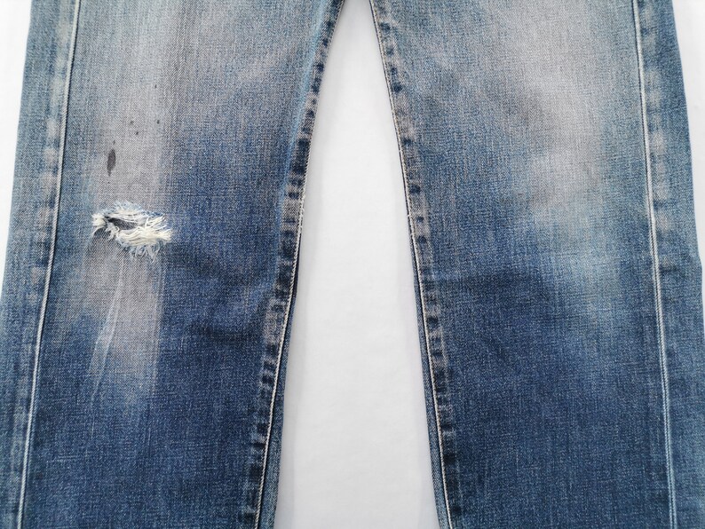 May include: Close-up of blue denim jeans with a distressed, worn appearance. The jeans have a ripped section on the left leg, showing frayed edges. Fading and discoloration are visible, adding to the vintage aesthetic.