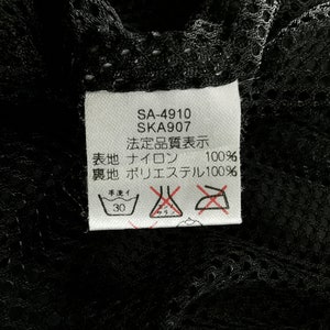 May include: A close-up of a white tag with black text and symbols, attached to a black mesh fabric. The tag includes the text "SA-4910", "SKA907", and Japanese characters. Washing instructions are also visible.