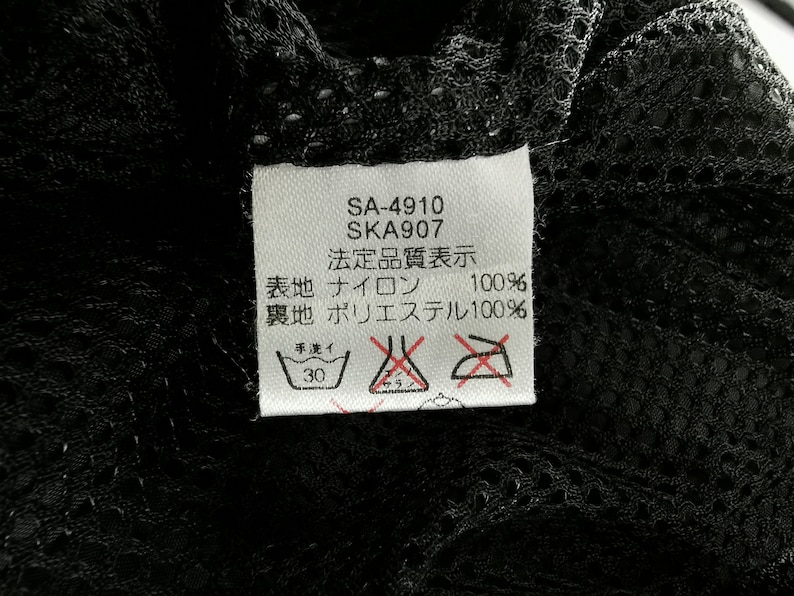 May include: A close-up of a white tag with black text and symbols, attached to a black mesh fabric. The tag includes the text "SA-4910", "SKA907", and Japanese characters. Washing instructions are also visible.