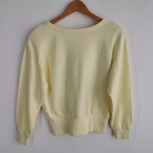 May include: A pale yellow sweatshirt with long sleeves, a crew neck, and ribbed cuffs and hem. The garment is hanging on a wooden hanger against a plain white wall. The sweatshirt is a solid color.