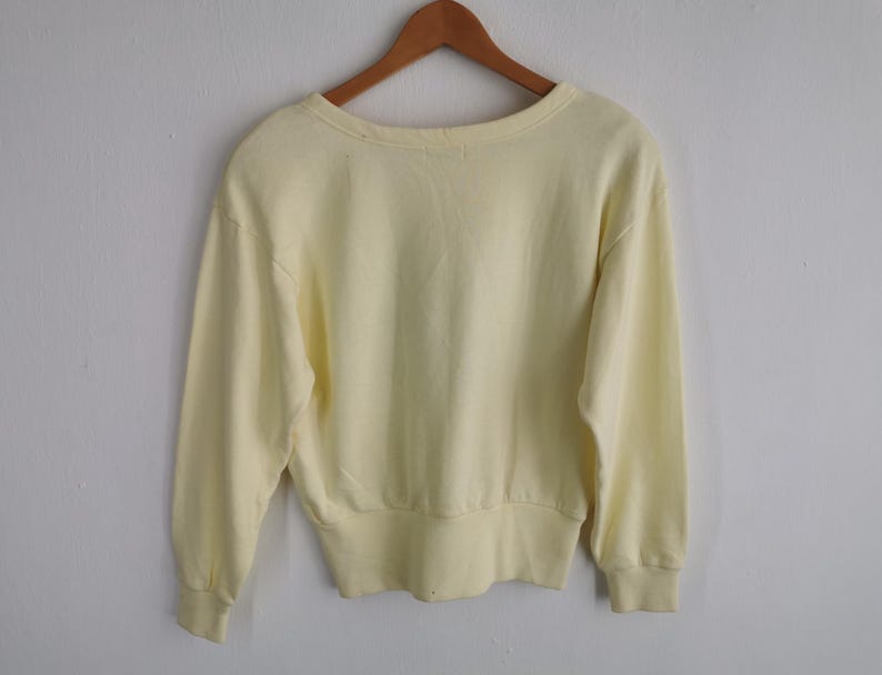 May include: A pale yellow sweatshirt with long sleeves, a crew neck, and ribbed cuffs and hem. The garment is hanging on a wooden hanger against a plain white wall. The sweatshirt is a solid color.