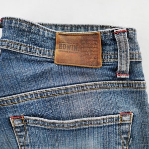 May include: Close-up of blue denim jeans with a brown leather patch. The patch features the brand name "EDWIN" and "GENUINE QUALITY" text. The jeans have yellow stitching and red accents.