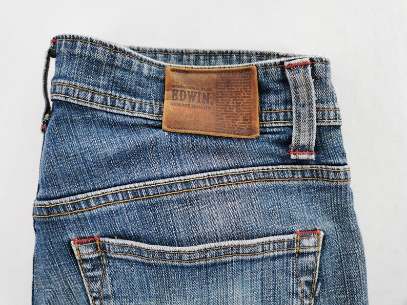 May include: Close-up of blue denim jeans with a brown leather patch. The patch features the brand name "EDWIN" and "GENUINE QUALITY" text. The jeans have yellow stitching and red accents.