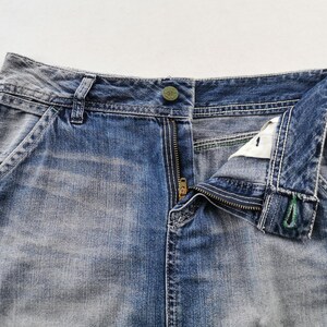 Pu&ograve; includere: Primo piano di jeans di denim blu con una cerniera in ottone parzialmente aperta. I jeans hanno una chiusura con bottone e dettagli di cucitura visibili. Il denim ha un aspetto sbiadito e usurato, con tasche e passanti per cintura.