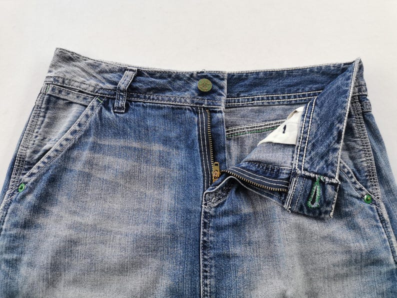 Pu&ograve; includere: Primo piano di jeans di denim blu con una cerniera in ottone parzialmente aperta. I jeans hanno una chiusura con bottone e dettagli di cucitura visibili. Il denim ha un aspetto sbiadito e usurato, con tasche e passanti per cintura.