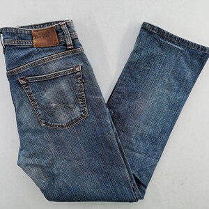 May include: Blue denim jeans with a faded wash. Features include a classic five-pocket design, a leather patch with the brand name, and a distinctive back pocket stitch. The jeans are folded, showcasing the details.