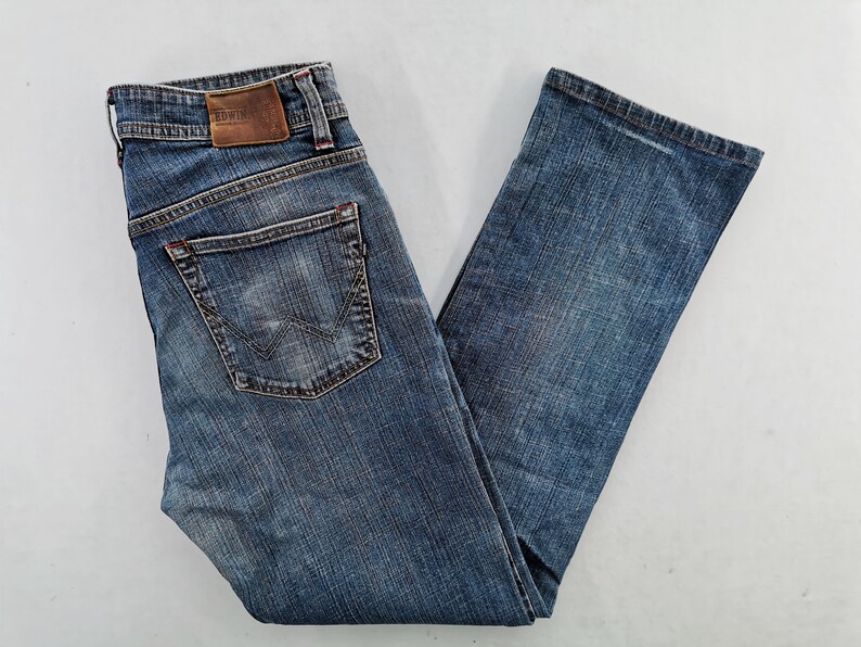 May include: Blue denim jeans with a faded wash. Features include a classic five-pocket design, a leather patch with the brand name, and a distinctive back pocket stitch. The jeans are folded, showcasing the details.