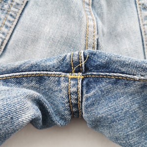 May include: Close-up of light blue denim jeans. The image highlights the stitching details, with yellow thread used to reinforce the seams and pockets. The fabric has a faded, worn appearance.