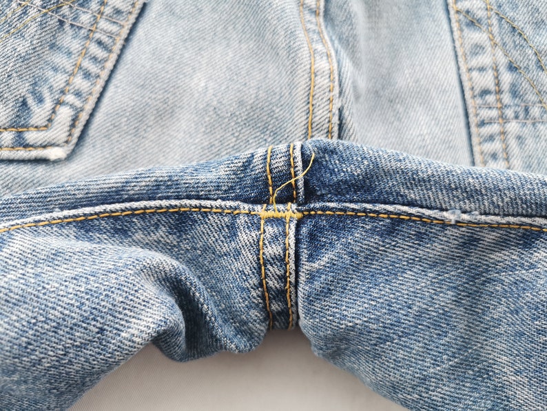 May include: Close-up of light blue denim jeans. The image highlights the stitching details, with yellow thread used to reinforce the seams and pockets. The fabric has a faded, worn appearance.