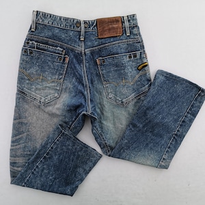 May include: Blue denim jeans with a faded wash. The jeans feature a classic five-pocket design, a brown leather patch, and decorative stitching on the back pockets. The jeans have a straight leg cut.