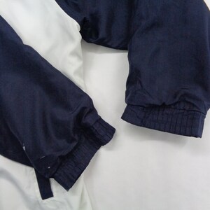 May include: A white and navy blue jacket with elastic cuffs. The jacket has a color block design with white on the upper body and navy blue on the sleeves. The sleeves are gathered at the wrist with elastic.